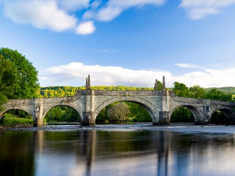 General Wade's Bridge, eine historische Steinbrücke mit fünf Bögen über einem Fluss in Aberfeldy. Üppige grüne Bäume rahmen beide Seiten unter blauem Himmel.