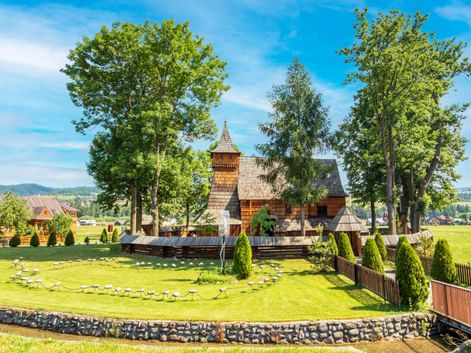 Traditionelle Holzkirche in Dąbno mit Schindeldach und Glockenturm, umgeben von hohen Bäumen und gepflegtem Rasen mit kreisförmiger Steinumrandung.