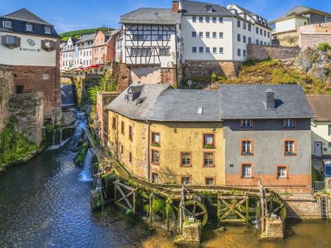Wasserfall fließt durch Saarburgs Altstadt mit traditionellen Fachwerkhäusern, Steinmauern und einer Holzbrücke über dem Bach.