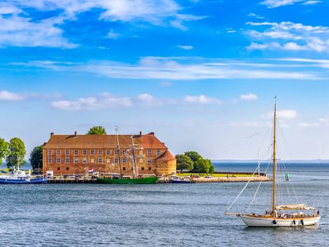 Schloss Sonderburg, ein großes Backsteingebäude mit orangefarbenem Ziegeldach, am Wasser. Ein Segelboot fährt im Vordergrund auf blauem Wasser.
