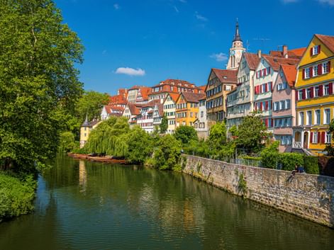 Reihe bunter Fachwerkhäuser mit roten Ziegeldächern am Neckar in Tübingen. Ein Kirchturm erhebt sich hinter den Gebäuden unter blauem Himmel.