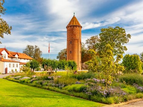 Roter Backsteinwasserturm in Praestø mit Kegeldach, umgeben von Gartenbeeten und grünem Rasen. Weißes Gebäude mit rotem Dach links sichtbar.