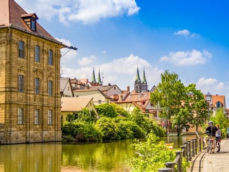 Radfahrer am Flussufer in der Bamberger Altstadt mit historischem Sandsteingebäude, bunten Häusern und Doppelkirchtürmen unter blauem Himmel.