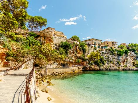 Malerische Bucht in Porto Cristo mit türkisfarbenem Wasser, Sandstrand und Küstenpromenade. Steingebäude und Hotels auf Felsenklippen mit Pinien.