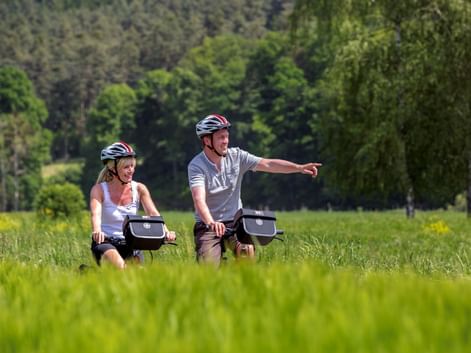Zwei Radfahrer mit Helmen fahren durch grüne Wiese im Altmühltal. Mann zeigt nach vorne, Wald im Hintergrund. Sonniger Sommertag.