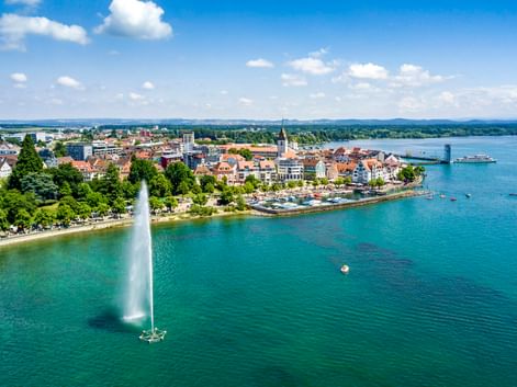 Luftaufnahme von Friedrichshafen am Bodensee mit hoher Fontäne im türkisfarbenen Wasser, historischer Altstadt, Kirchturm und Fahrgastschiff.