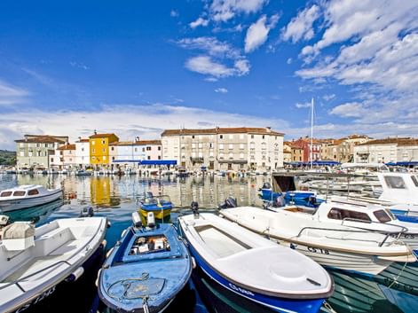 Hafen auf der Insel Cres mit bunten Booten im ruhigen Wasser. Mediterrane Gebäude säumen die Uferpromenade unter blauem Himmel mit weißen Wolken.