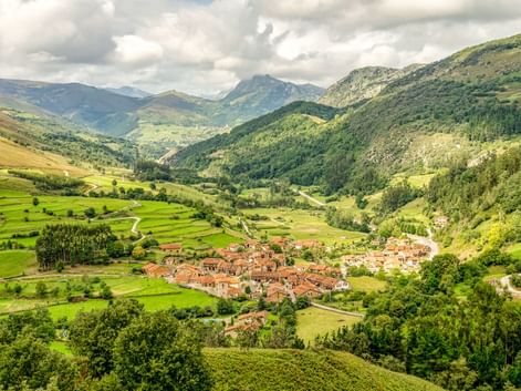 Luftaufnahme eines Dorfes mit Terrakottadächern in einem grünen Tal, umgeben von bewaldeten Bergen unter bewölktem Himmel in Spanien.