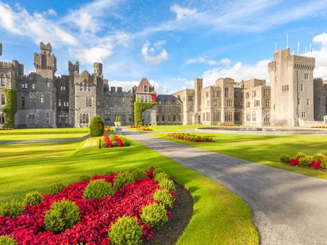 Ashford Castle in Irland mit Steintürmen und Türmchen. Gepflegte Rasenflächen mit roten und grünen Blumenbeeten säumen die Auffahrt unter blauem Himmel.