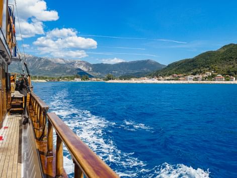 Blick vom Holzbootsdeck auf blaues Wasser nahe Lefkada. Berge und Küstenstadt in der Ferne unter klarem Himmel sichtbar.