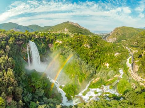 Luftaufnahme des Wasserfalls Cascata delle Marmore in Italien mit Regenbogen im Nebel, umgeben von grünen Bergen und Serpentinenstraße.