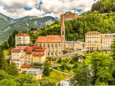Pfarrkirche mit hohem Turm und rotem Dach in Bad Gastein, umgeben von bunten Gebäuden am Hang mit bewaldeten Bergen im Hintergrund.