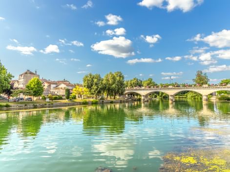 Charente-Fluss in Cognac mit ruhigem grünen Wasser, das Wolken spiegelt. Steinbrücke mit mehreren Bögen überspannt den Fluss, gesäumt von Bäumen.