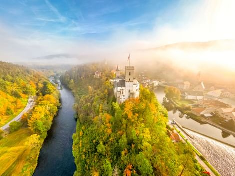 Luftaufnahme der Burg Rosenberg auf einem bewaldeten Hügel, umgeben von einer Flussschleife. Herbstbäume in goldenen Farben, Morgennebel und Sonnenlicht.