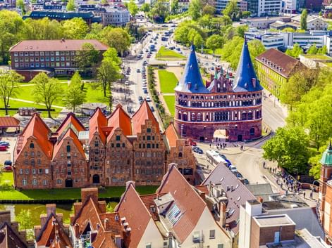 Luftaufnahme von Lübeck mit dem ikonischen Holstentor mit Zwillingstürmen und roten Backsteingebäuden, umgeben von grünen Parks und Stadtgebieten.