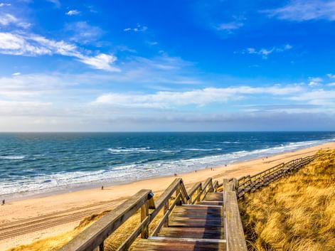 Holztreppe führt durch Dünengras zum Sandstrand in Wenningstedt, Sylt. Blauer Himmel mit weißen Wolken über der Nordsee.