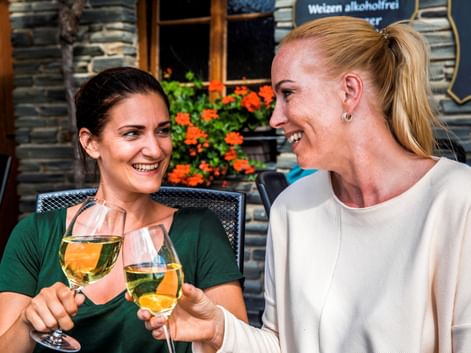 Zwei lächelnde Frauen stoßen mit Weißweingläsern auf einer Außenterrasse an. Orangefarbene Blumen und Weinfässer im Hintergrund.