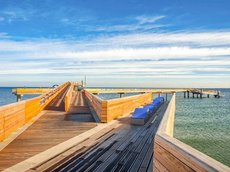 Moderne Holzsteganlage mit blauen Bänken, die in die Ostsee in Heiligenhafen hineinragt, unter blauem Himmel mit vereinzelten Wolken.