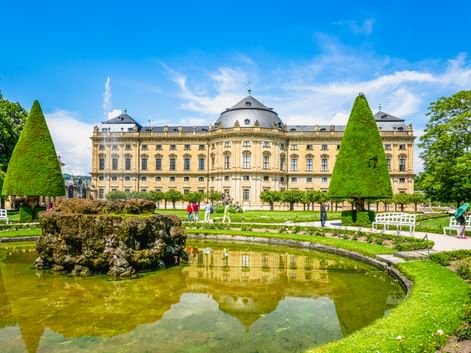 Würzburger Residenz mit gelber Barockfassade und Kuppel, vom formalen Garten mit Zierteich und kegelförmigen Bäumen aus gesehen.