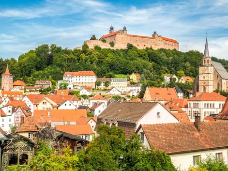 Plassenburg auf einem Hügel über der Stadt Kulmbach mit rotgedeckten Häusern, Kirchturm und grünen Bäumen unter blauem Himmel in Franken.