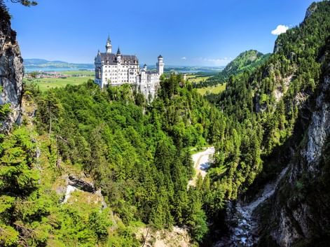 Schloss Neuschwanstein thront auf einem Hügel, umgeben von dichtem Wald und Bergen. Eine Schlucht mit Felsklippen rahmt die Ansicht in Bayern ein.