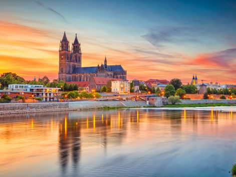 Magdeburger Dom mit Doppeltürmen an der Elbe bei Sonnenuntergang. Farbenfroher Himmel spiegelt sich im ruhigen Wasser, Stadtgebäude sichtbar.