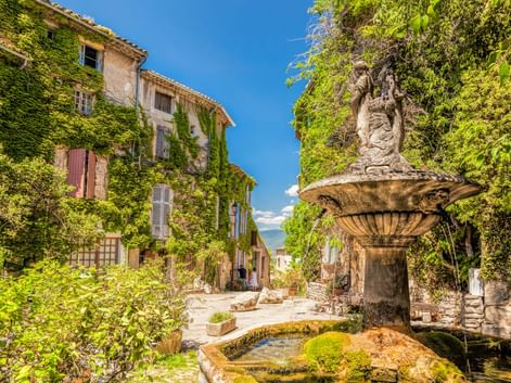 Steinbrunnen mit Statue auf dem Dorfplatz von Saignon, umgeben von efeubedeckten Gebäuden und üppigem Grün unter blauem Himmel.