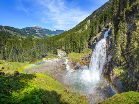 Krimmler Wasserfälle stürzen über Felsklippen mit Regenbogen im Nebel, umgeben von grünen Almwiesen und bewaldeten Bergen in Österreich.