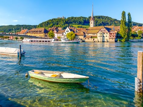 Blick auf Stein am Rhein am Rheinufer mit Booten im Vordergrund, historischen Gebäuden, Kirchturm und bewaldeten Hügeln im Hintergrund.