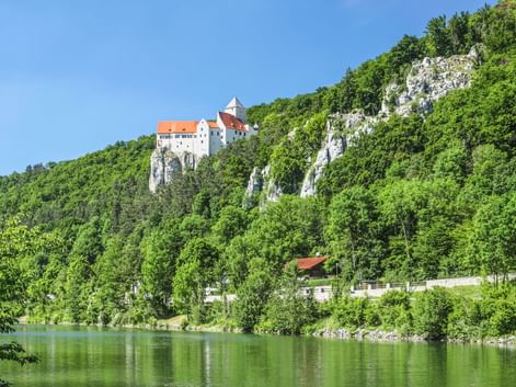 Burg Prunn thront auf weißen Kalksteinfelsen über grünen Wäldern bei Riedenburg. Die Altmühl fließt im Vordergrund.