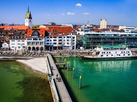 Aerial view of Friedrichshafen harbor with a ferry boat docked at the pier, green lake water, promenade with people, and historic buildings with a church tower.