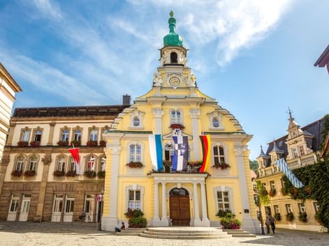 Gelbes barockes Rathaus in Kulmbach mit grünem Turm und Uhr. Bayerische und deutsche Flaggen hängen an der Fassade. Historische Gebäude rahmen den Platz.