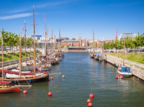Traditionelle hölzerne Segelboote liegen in einem Kanal im Kieler Hafen unter blauem Himmel, moderne Gebäude und grüne Bäume säumen das Ufer.
