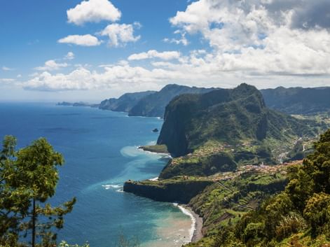 Dramatische Küstenklippen bei Funchal, Madeira, mit grüner Vegetation, türkisfarbenem Ozean und kleinen Stränden unter blauem Himmel mit Wolken.