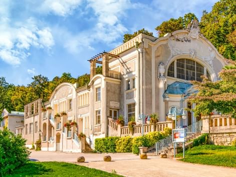 Eleganter Jugendstil-Pavillon im Kurpark Baden bei Wien mit verzierter Fassade, Bogenfenstern und großer Treppe, umgeben von grünen Rasenflächen.