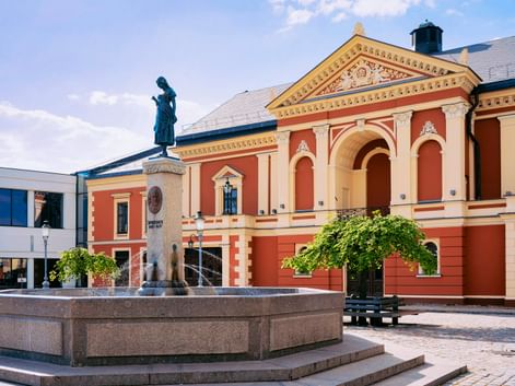 Historisches Klaipeda Theater in Korallen- und Cremefarben mit klassischer Architektur. Eine Bronzestatue steht auf einem Brunnen auf dem Platz.