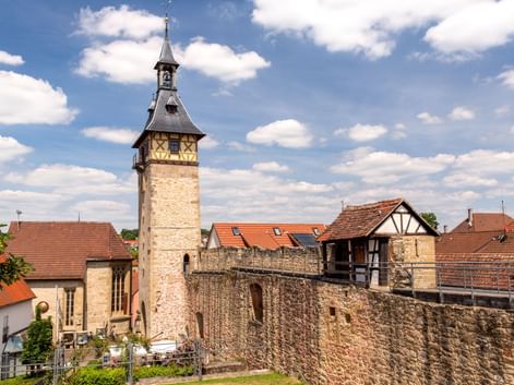 Stone tower with pointed roof and clock rising above old town wall in Marbach. Half-timbered houses with red tile roofs visible behind the fortification.