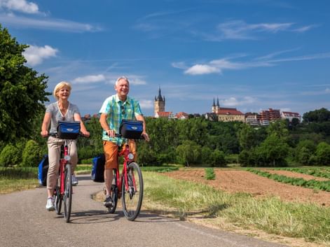 Zwei Radfahrer mit roten Rädern auf einem asphaltierten Weg durch Felder, mit Bad Wimpfens historischer Skyline und Kirchtürmen im Hintergrund.