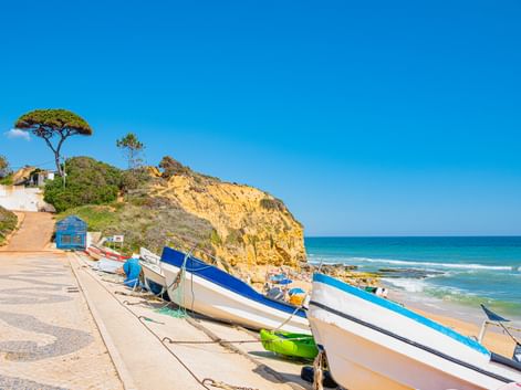 Bunte Fischerboote auf einer Promenade in Olhos de Água, mit goldenen Klippen, Pinien und türkisfarbenem Meer unter blauem Himmel.