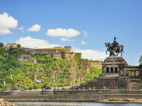 Equestrian monument at Deutsches Eck in Koblenz with fortress Ehrenbreitstein on hillside. International flags line the riverside promenade.