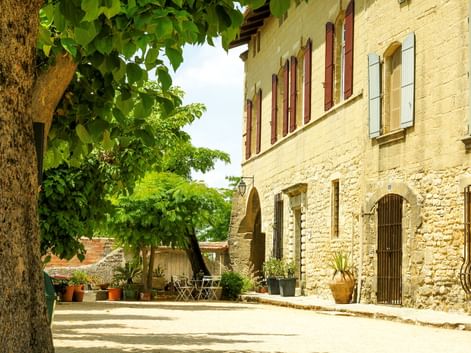 Traditionelles Steinhaus bei Uzès mit Bogentüren und Fensterläden. Ein großer Baum spendet Schatten über einem sonnigen Innenhof mit Topfpflanzen.