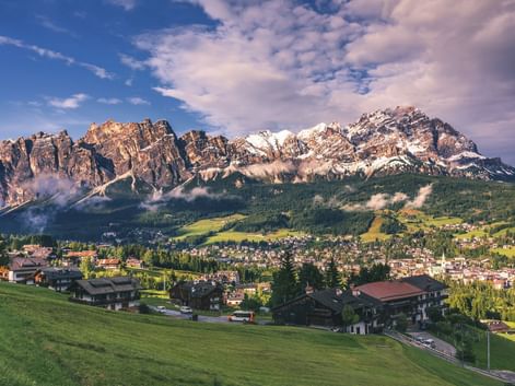 Alpendorf in Südtirol mit traditionellen Häusern auf grünen Wiesen. Dramatische Dolomitengipfel erheben sich hinter Wäldern unter blauem Himmel.