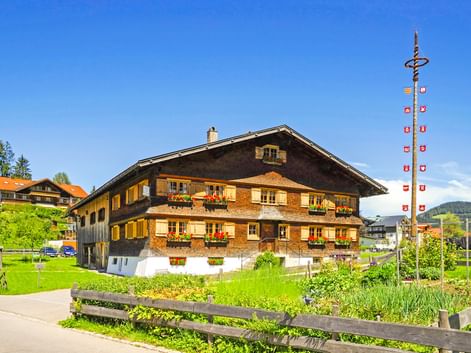 Traditionelles bayerisches Holzhaus mit Blumenkästen in Oberstaufen. Ein dekorativer Maibaum mit Fahnen steht neben dem Gebäude auf grüner Wiese.