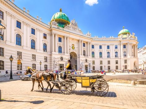 Gelber Fiaker vor der Alten Hofburg in Wien mit weißer Barockfassade und grünen Kupferkuppeln unter blauem Himmel.