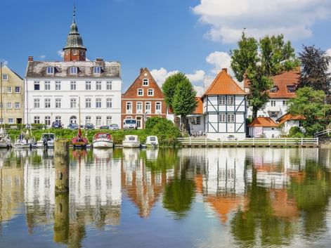 Historischer Hafen von Glückstadt mit bunten Gebäuden, die sich im ruhigen Wasser spiegeln. Weißes Gebäude mit Turm, Fachwerkhäuser und Boote am Kai.
