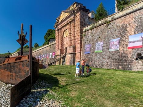 Historisches Festungstor in Neuf-Brisach mit roter Sandsteinfassade und Backsteinmauern. Radfahrer stehen auf grünem Rasen neben rostiger Metallskulptur unter blauem Himmel.