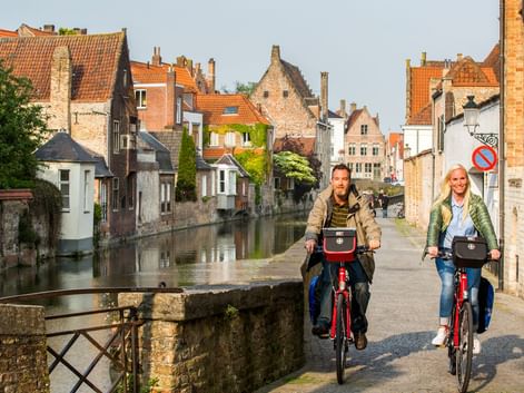 Zwei Radfahrer auf roten Fahrrädern auf einer Kopfsteinpflasterstraße neben einem Kanal in Flandern, mit historischen Backsteingebäuden und orangefarbenen Ziegeldächern.