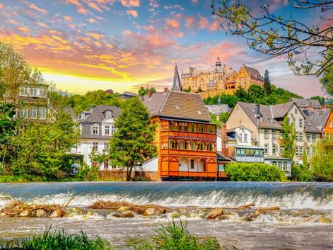 Marburg cityscape with colorful historic buildings along a weir on the Lahn River. Marburg Castle sits on the hilltop under a dramatic sunset sky.