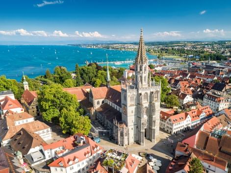 Luftaufnahme des Konstanzer Münsters mit gotischem Turm über den roten Dächern der Altstadt und dem Bodensee mit Segelbooten unter blauem Himmel.