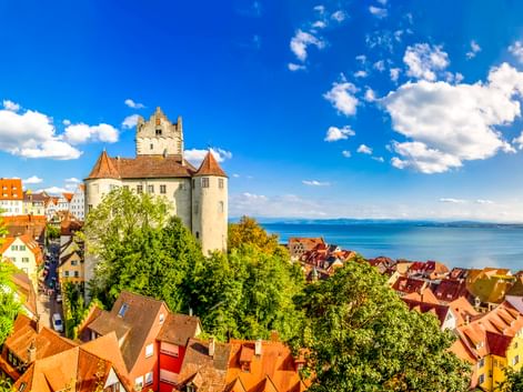 Burg Meersburg mit weißen Mauern und Turm über rotgedeckten Häusern, mit Blick auf den Bodensee unter blauem Himmel mit weißen Wolken.